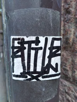 Close-up view of a sticker featuring an abstract design in black on a white background. The sticker is affixed to a dark brown pole. The design appears to be hand-drawn or painted, showing thick black lines and shapes that resemble stylized characters or symbols. The sticker shows signs of wear and creasing.