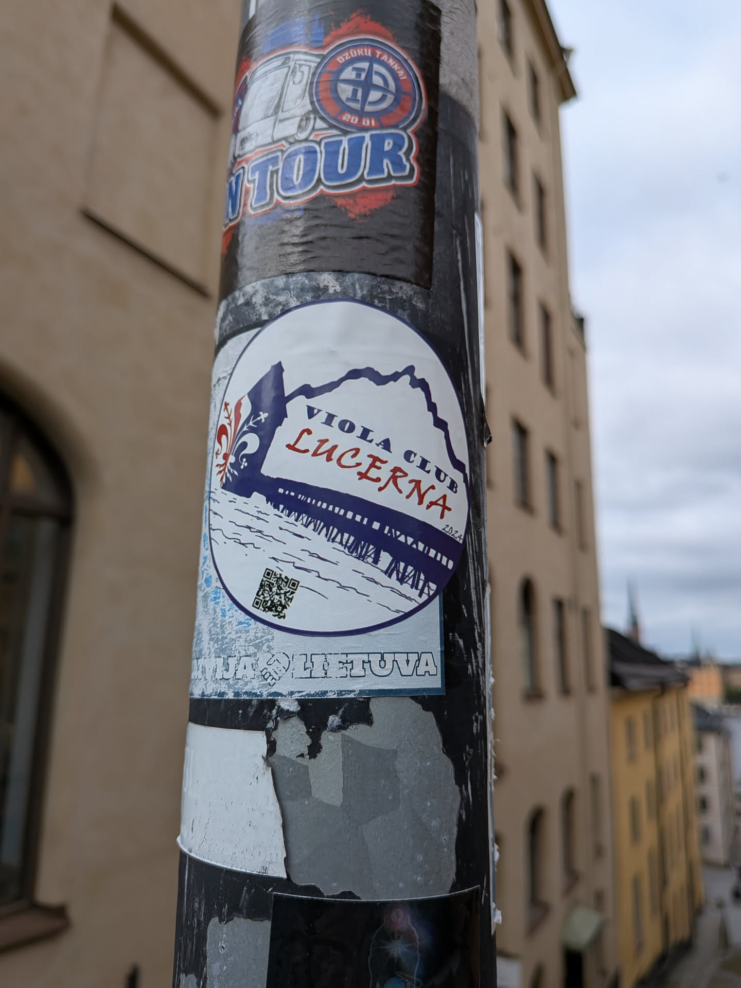 The sticker is circular with a white background. At the top left, it features a stylized fleur-de-lis in dark blue. The text "VIOLA CLUB" is written in dark blue uppercase letters, and "LUCERNA" is written below it in bold red uppercase letters. Below the text, there is a depiction of a pier or bridge with railings, rendered in dark blue lines against a white background, with a blue mountain range in the distance. A small QR code is positioned on the left side of the pier. The year "2024" is written in small red text to the right of the pier. A thin blue border encircles the sticker. The bottom of the sticker has some blue and white distressed elements and text which is partially obscured.