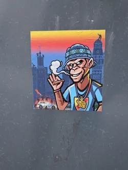 The sticker features a cartoon-style monkey character with brown fur, a wide grin, and a mischievous look. It wears a blue bucket hat adorned with green cannabis leaves and a light blue t-shirt with dark blue stripes. Around its neck is a gold chain with a large 'DIF' pendant in gold, red, and yellow. The monkey is holding a lit cigar or joint, from which a puff of white smoke emanates. On its right forearm, a tattoo reads 'RUDE BOYS'. The background depicts a stylized urban cityscape with tall buildings under a gradient sky transitioning from yellow to orange to red and then blue. In the lower left, a white and blue police car with 'GRIS' written on its side is visible, with small figures and a red flare in the foreground.