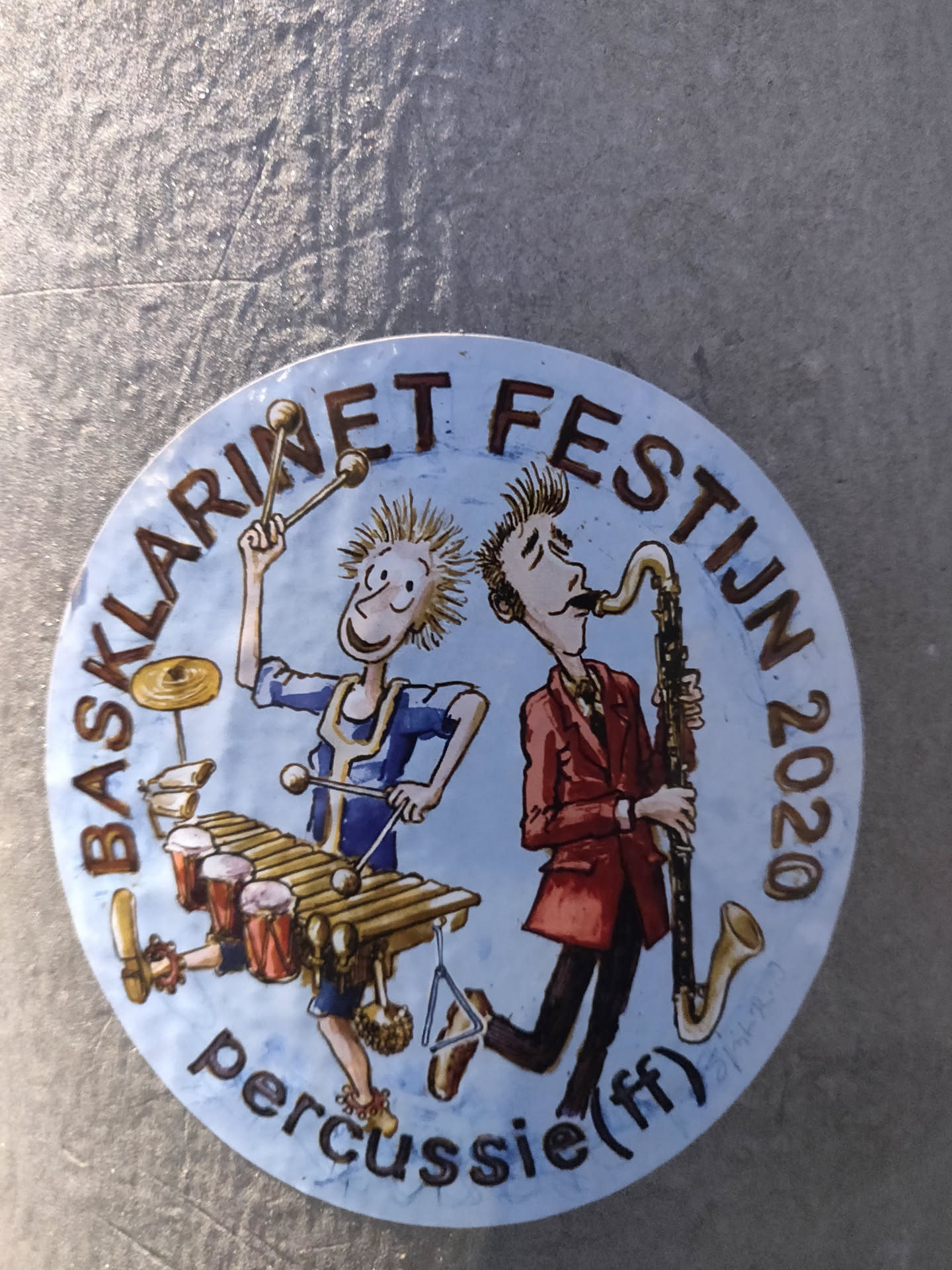 Round sticker featuring an illustration of two cartoon musicians; one playing the xylophone and the other a bass clarinet. The text around the illustration says "Bassklarinet Festijn 2020" and  "percussie (f)" at the bottom. 