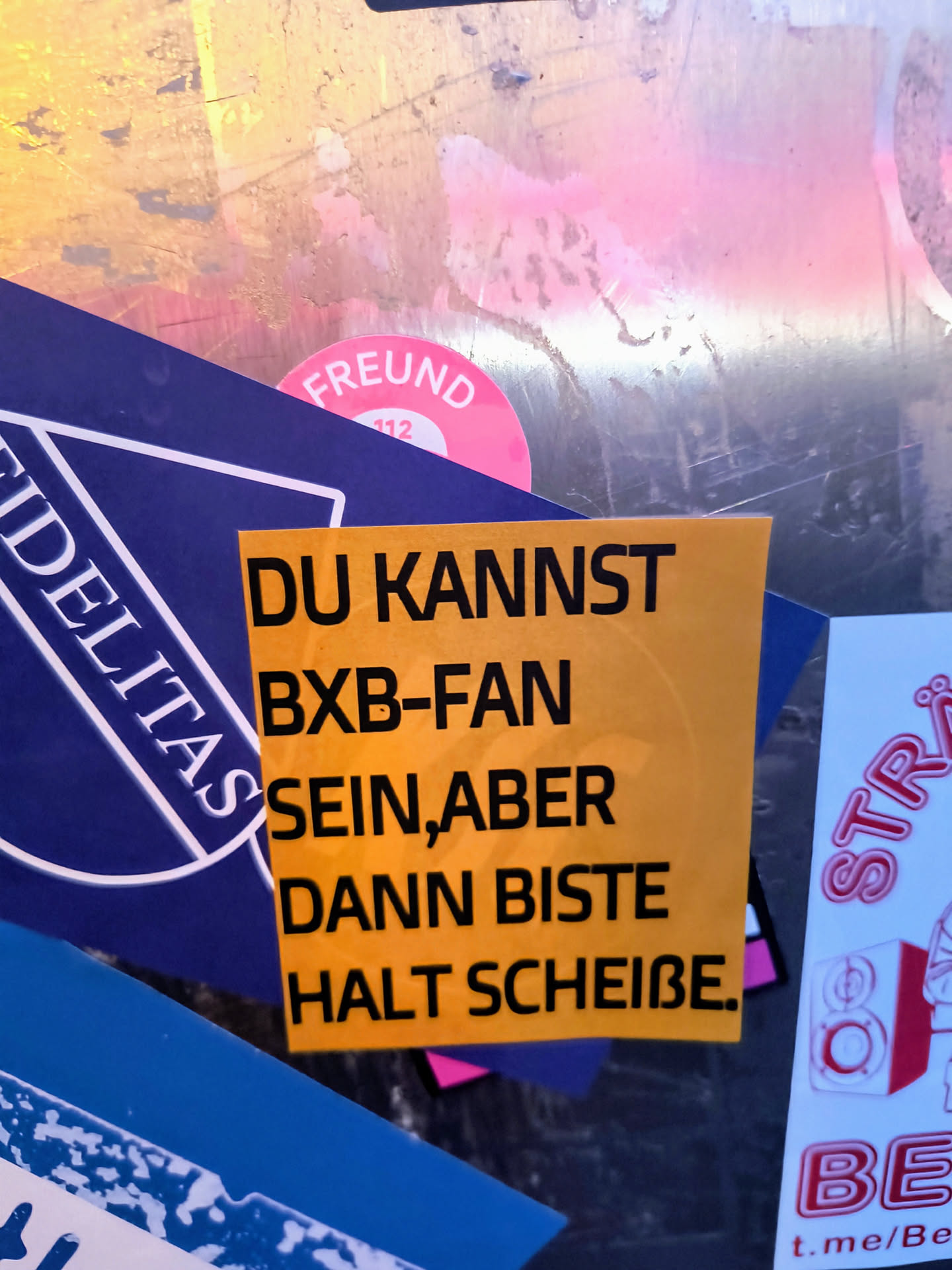 A yellow rectangular sticker with the text "DU KANNST BXB-FAN SEIN, ABER DANN BISTE HALT SCHEIßE." in bold black letters. The sticker is stuck on a metallic surface along with other stickers.