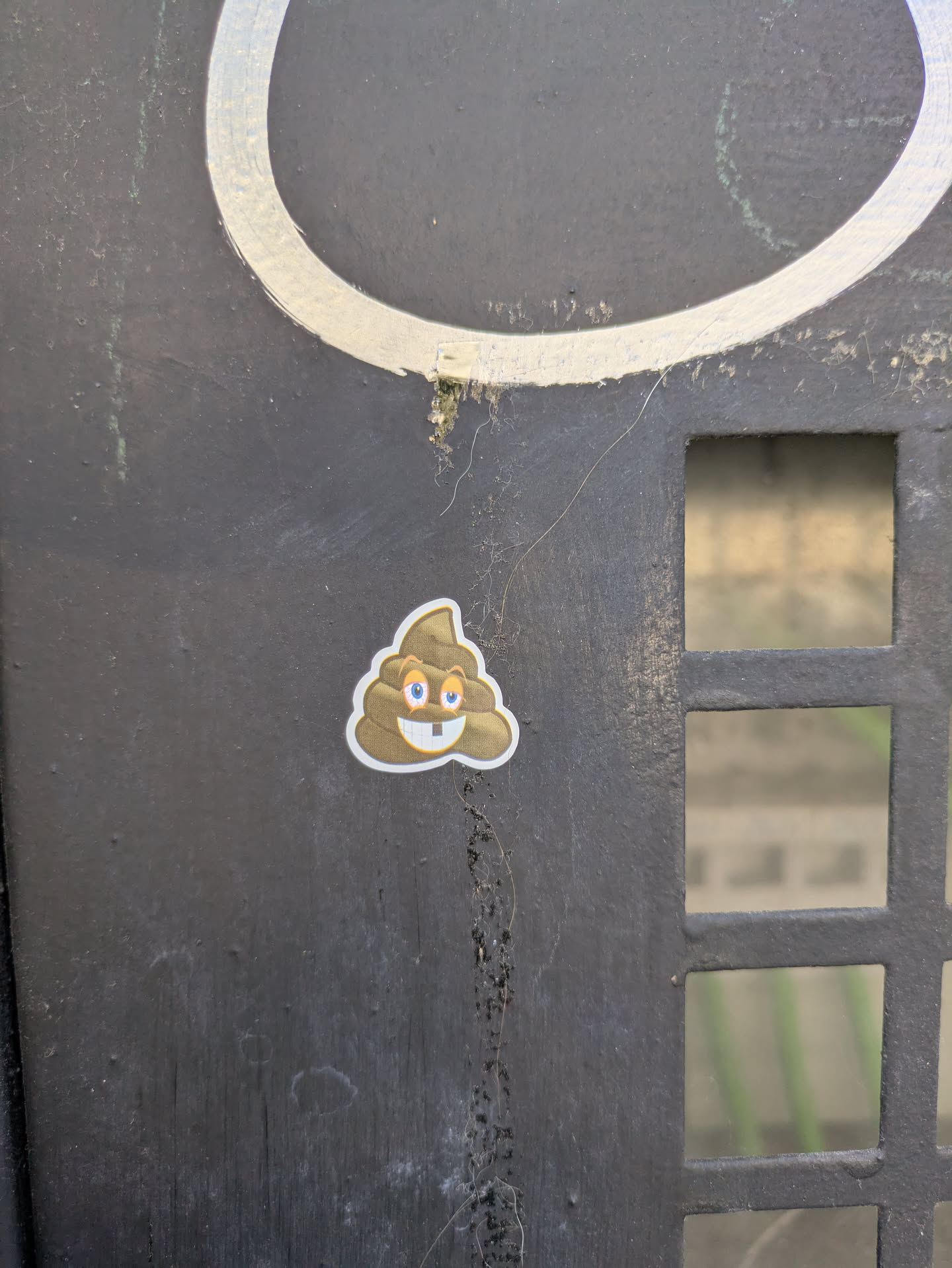 A poop emoji sticker is stuck on a black metal surface. The poop emoji is cartoonish, with big blue eyes and two buck teeth.