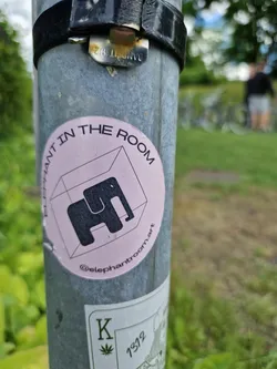 A pink and black sticker with the text "Elephant in the room" and an illustration of an elephant inside a cube. The sticker also includes the Instagram handle @elephantroom.art
