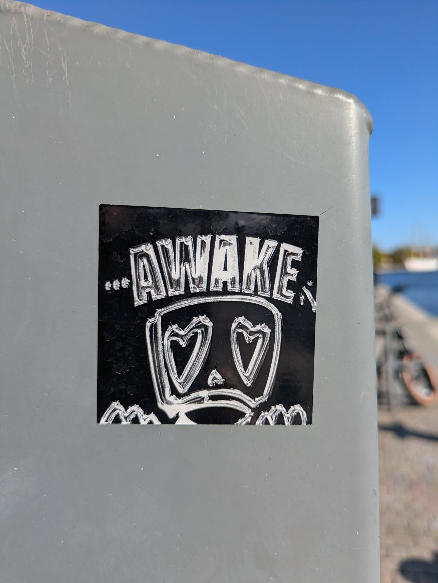 A black sticker with white outlines, featuring the word "AWAKE" in a stylistic font at the top. Below, there is a character with heart-shaped eyes and a sad mouth, evoking a sense of melancholy or love-struck emotion. The artwork has a distinct, hand-drawn feel.
