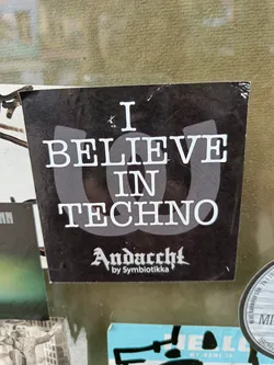 Black and white sticker with the text "I Believe in Techno" and the logo "Andacht by Symbiotikka".