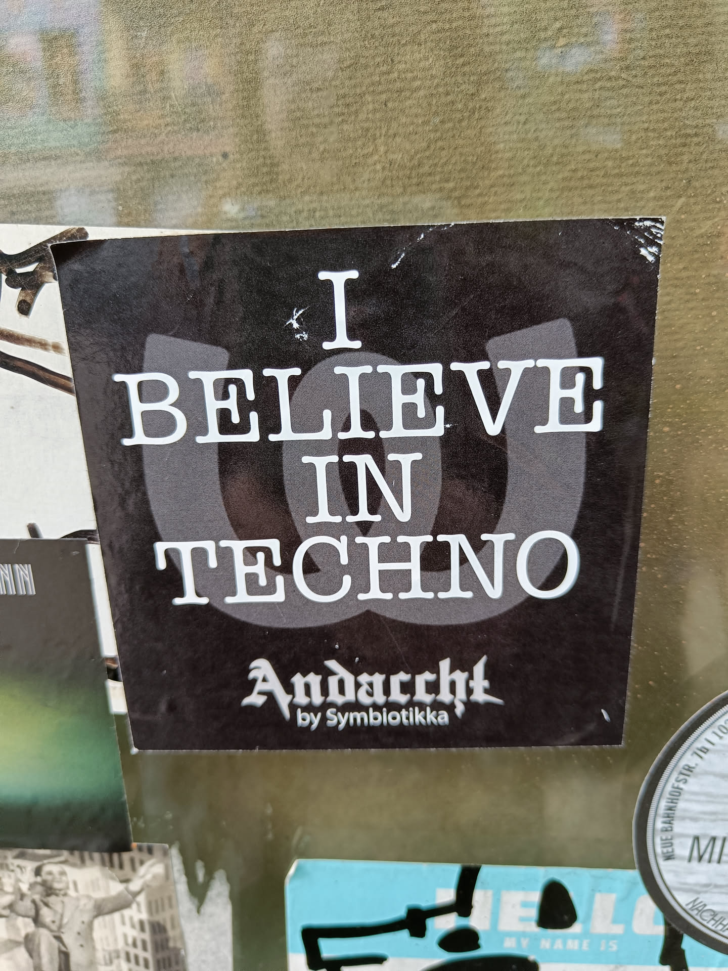 Black and white sticker with the text "I Believe in Techno" and the logo "Andacht by Symbiotikka".
