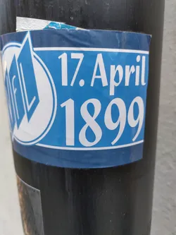 A blue sticker with the text "17. April 1899" and a logo. The logo appears to be a stylized "FL" inside a circle.