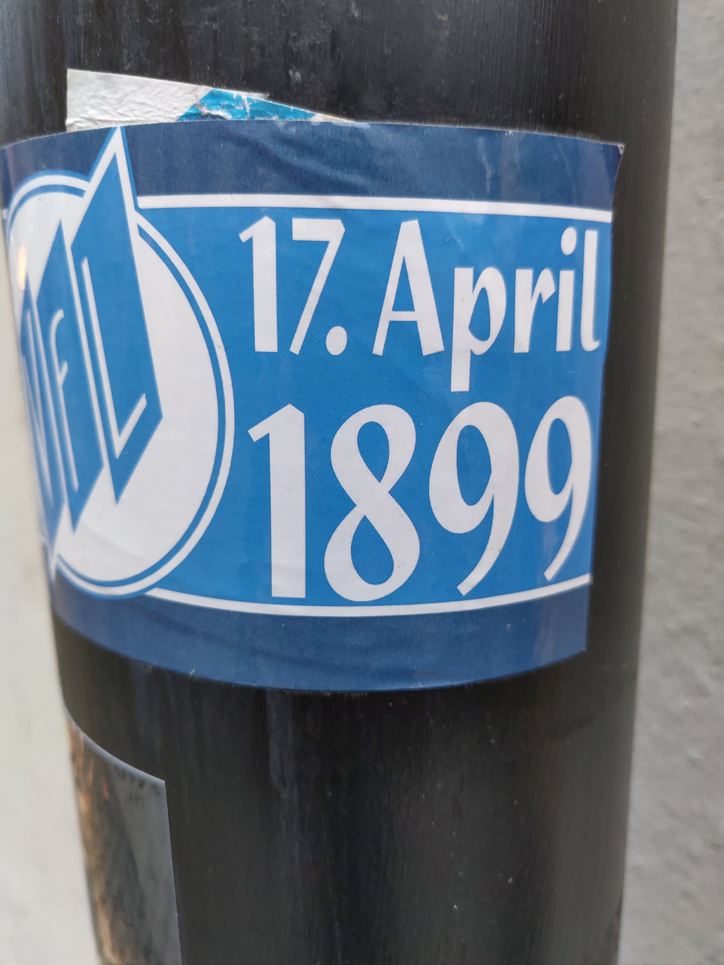 A blue sticker with the text "17. April 1899" and a logo. The logo appears to be a stylized "FL" inside a circle.