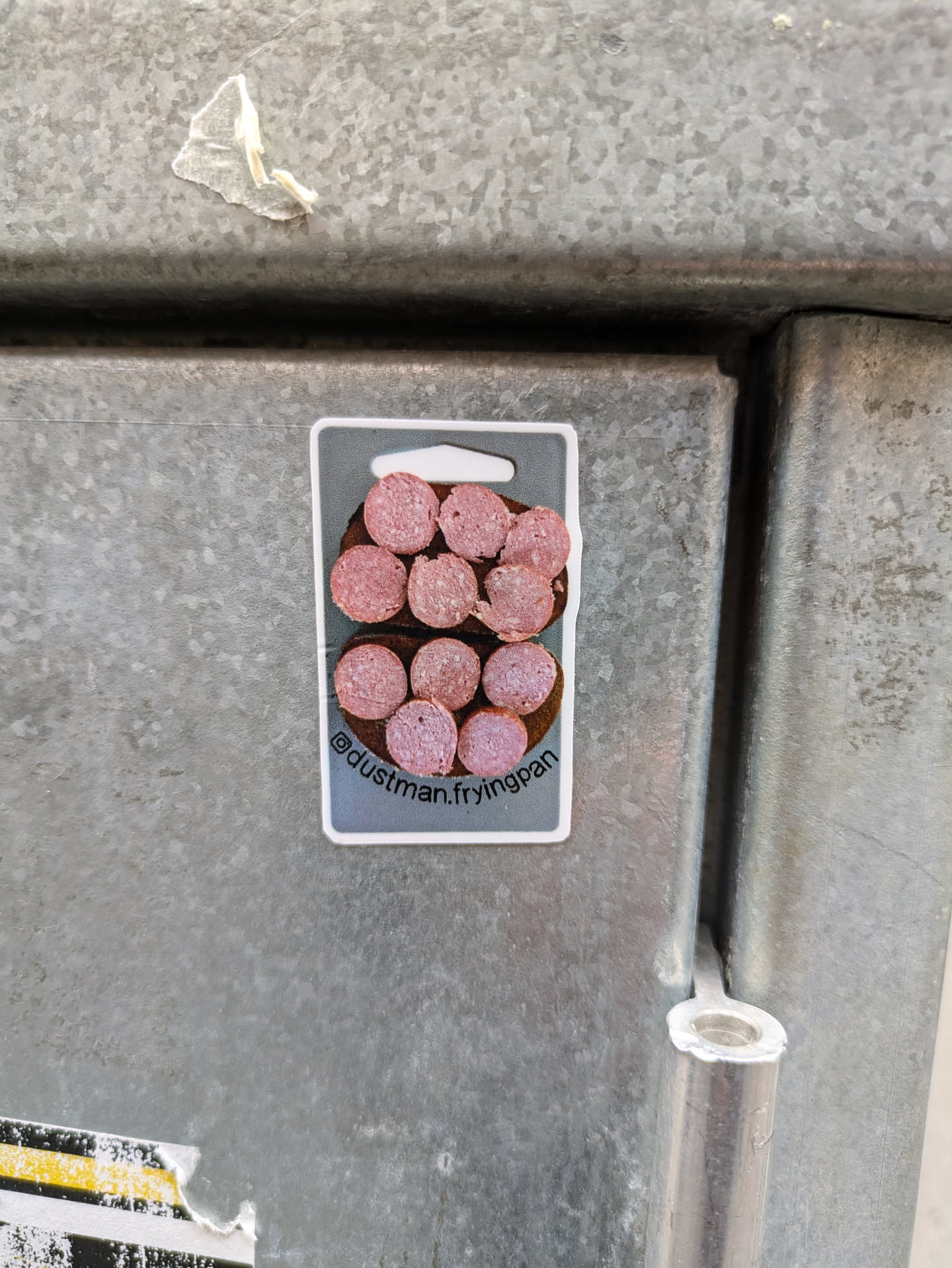 A sticker featuring a graphic of sliced sausages arranged on a frying pan. The sticker is rectangular with rounded corners and has a hang hole at the top. The text '@dustman.fryingpan' is printed at the bottom of the sticker.