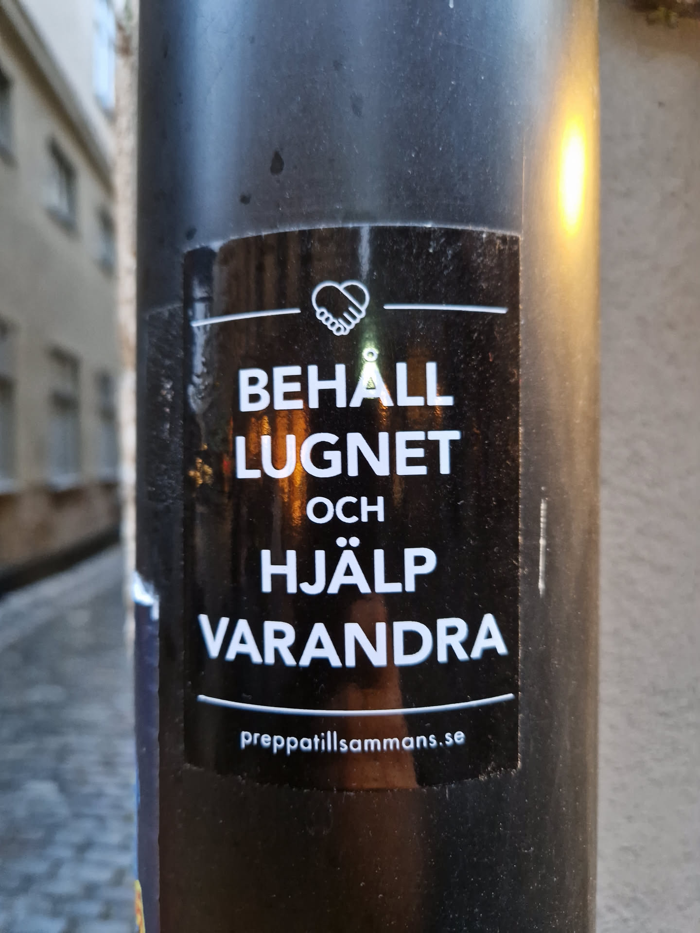 A black sticker with white text on a dark pole. The text translates to: "Keep calm and help each other."  A website address is also included at the bottom.
