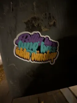 Street sticker A die-cut sticker showing three stacked lines of stylized bubble-script text with 3D block shading. The top line is rendered in purple ('Billig lyx'), the middle line in cyan-teal ('tung bas'), and the bottom line in warm yellow ('äkta vänskap'). The design is outlined with a white die-cut border and adhered to a dark, textured surface.