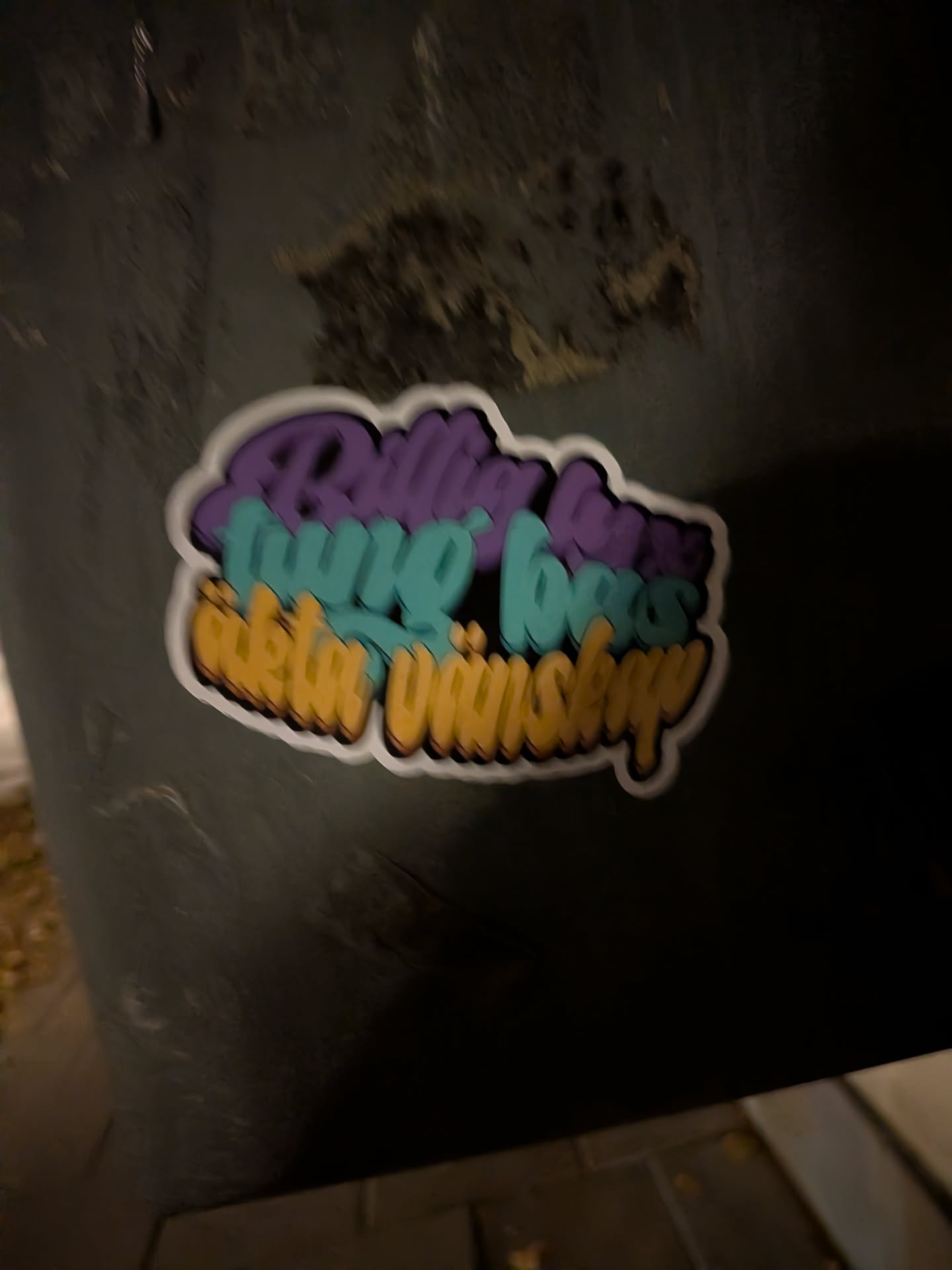 A die-cut sticker showing three stacked lines of stylized bubble-script text with 3D block shading. The top line is rendered in purple ('Billig lyx'), the middle line in cyan-teal ('tung bas'), and the bottom line in warm yellow ('äkta vänskap'). The design is outlined with a white die-cut border and adhered to a dark, textured surface.