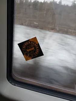 A square-shaped sticker, predominantly black with metallic gold or orange details, featuring a circular crest design. The crest includes a central shield-like emblem topped with a crown. Inside the shield, the letters 'A.I.K.' are prominently displayed, with '1891' beneath them. The text 'NORRA STAPLATS' is arched along the top inner edge of the circular border. The design also incorporates checkered patterns on the sides of the central shield and a star at the bottom. The overall aesthetic is heraldic and vintage, with a slight corner of the sticker appearing to peel from the window surface.