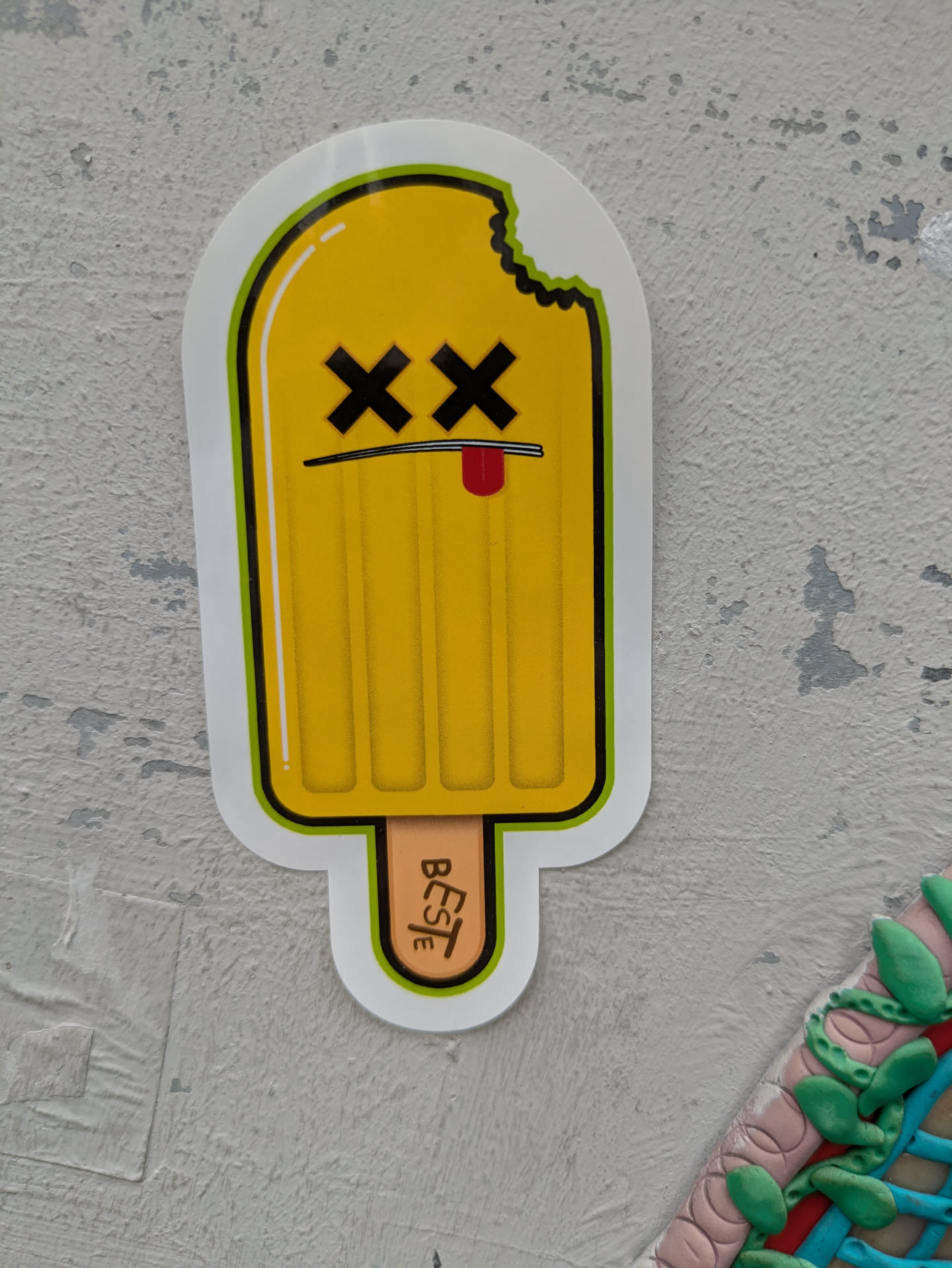 This vibrant sticker portrays a yellow popsicle with a distinct personality. It has a cartoonish face featuring black 'X's for eyes and a small red tongue peeking out from a thin black line that serves as a mouth, giving it a playful yet 'dead' or 'finished' appearance. A bite mark is visible on the upper right side of the popsicle. The light brown popsicle stick has the word "BESTe" written vertically in black, likely serving as an artist's signature or brand. The entire design is outlined in black and a bright lime green, and is placed on a light grey, textured wall.
