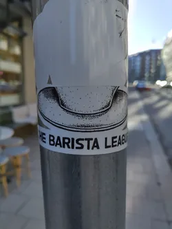 A black and white sticker featuring a drawing of a coffee cup and the text "The Barista League". The sticker is affixed to a metal pole outdoors.