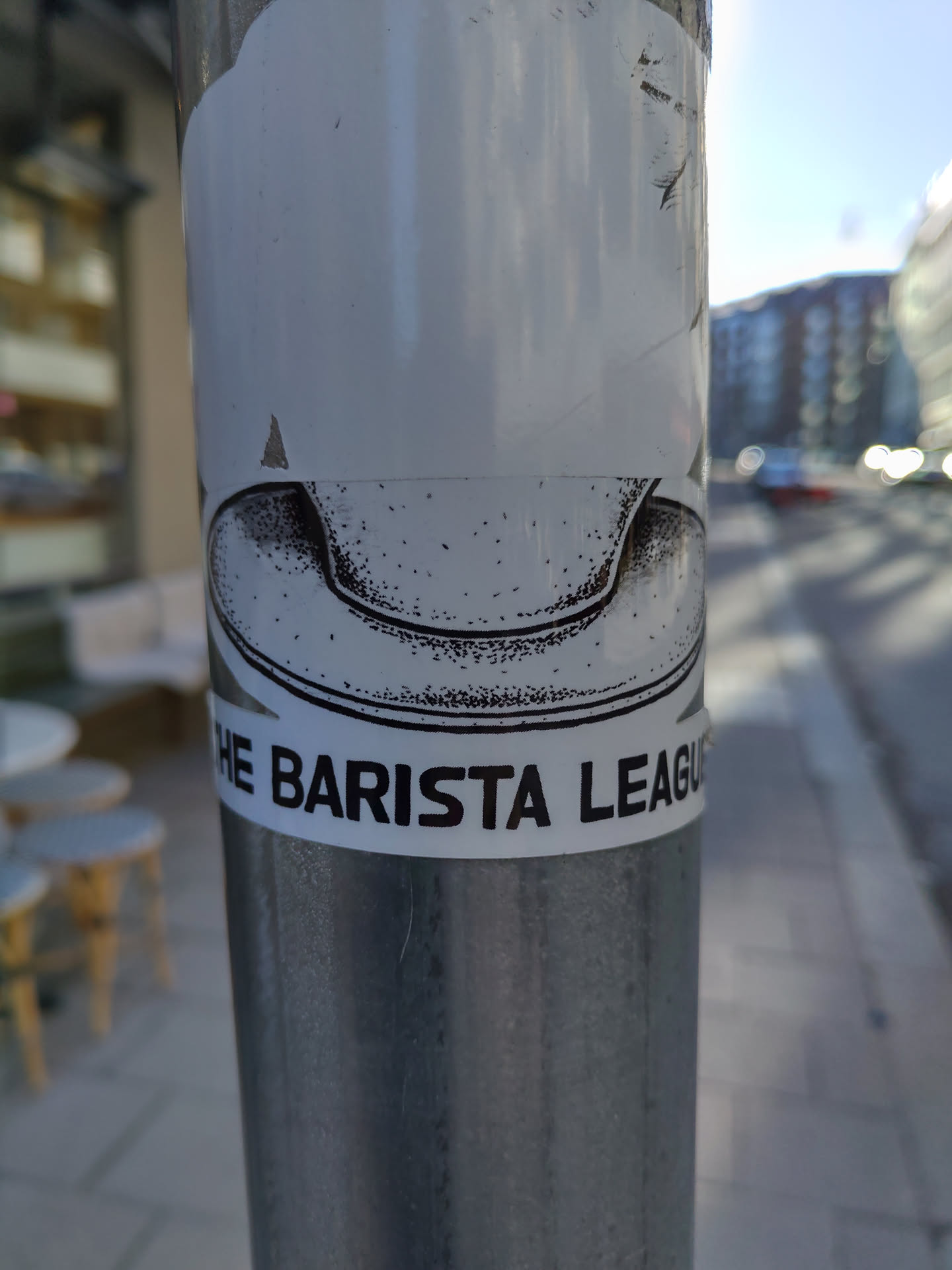 A black and white sticker featuring a drawing of a coffee cup and the text "The Barista League". The sticker is affixed to a metal pole outdoors.