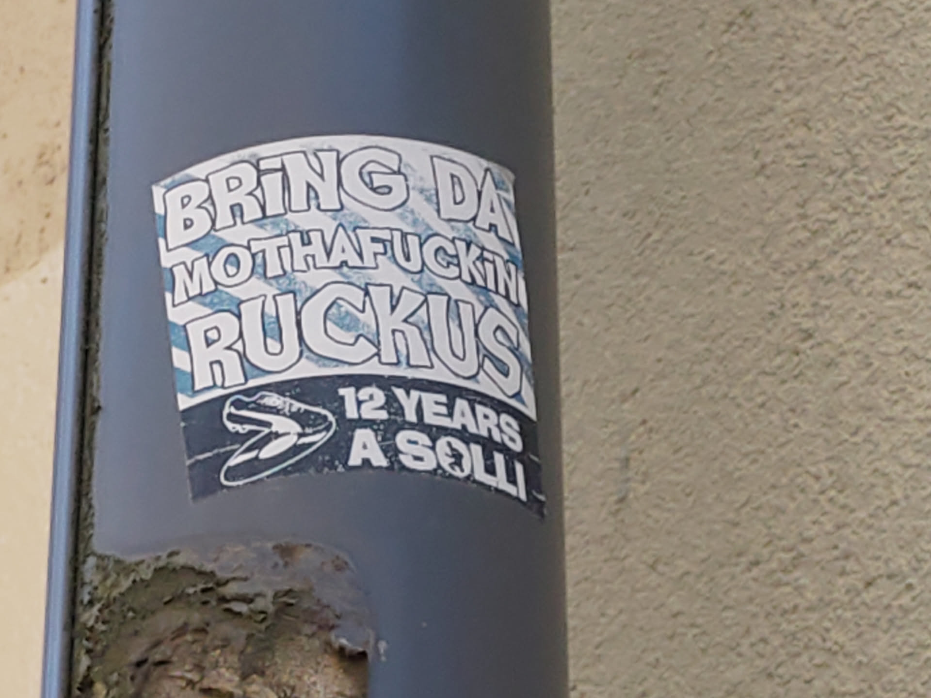 A sticker on a grey pipe with the text "Bring da motherfuckin ruckus" and a picture of a shoe. At the bottom it says "12 years a solli"