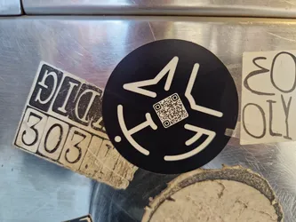A black circular sticker with a minimalist geometric design. The design features angular shapes and lines creating a stylized symbol. A QR code is incorporated into the center of the design. The sticker is affixed to a metallic surface, and there are other stickers visible around it.