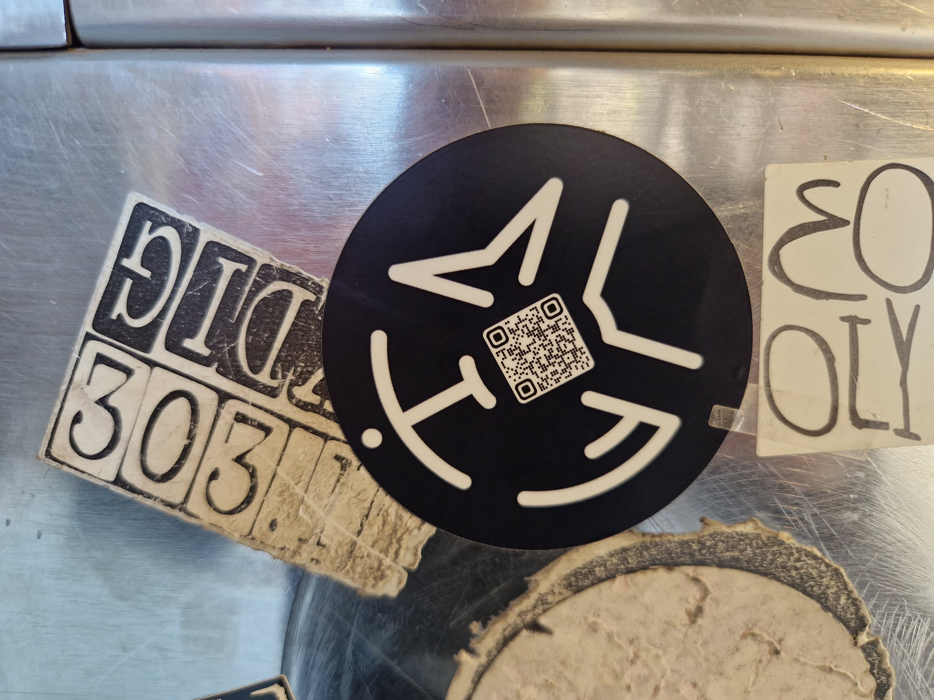 A black circular sticker with a minimalist geometric design. The design features angular shapes and lines creating a stylized symbol. A QR code is incorporated into the center of the design. The sticker is affixed to a metallic surface, and there are other stickers visible around it.