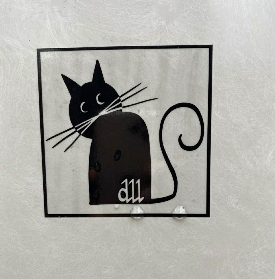 A square-shaped sticker featuring a stylized black cat with a whimsical tail.  The cat is depicted in a minimalist style, with simple lines and shapes. The sticker appears to be made of a clear vinyl material, and there are a few water droplets visible on its surface. The word "all" is written in a stylized font on the lower part of the cat's body.