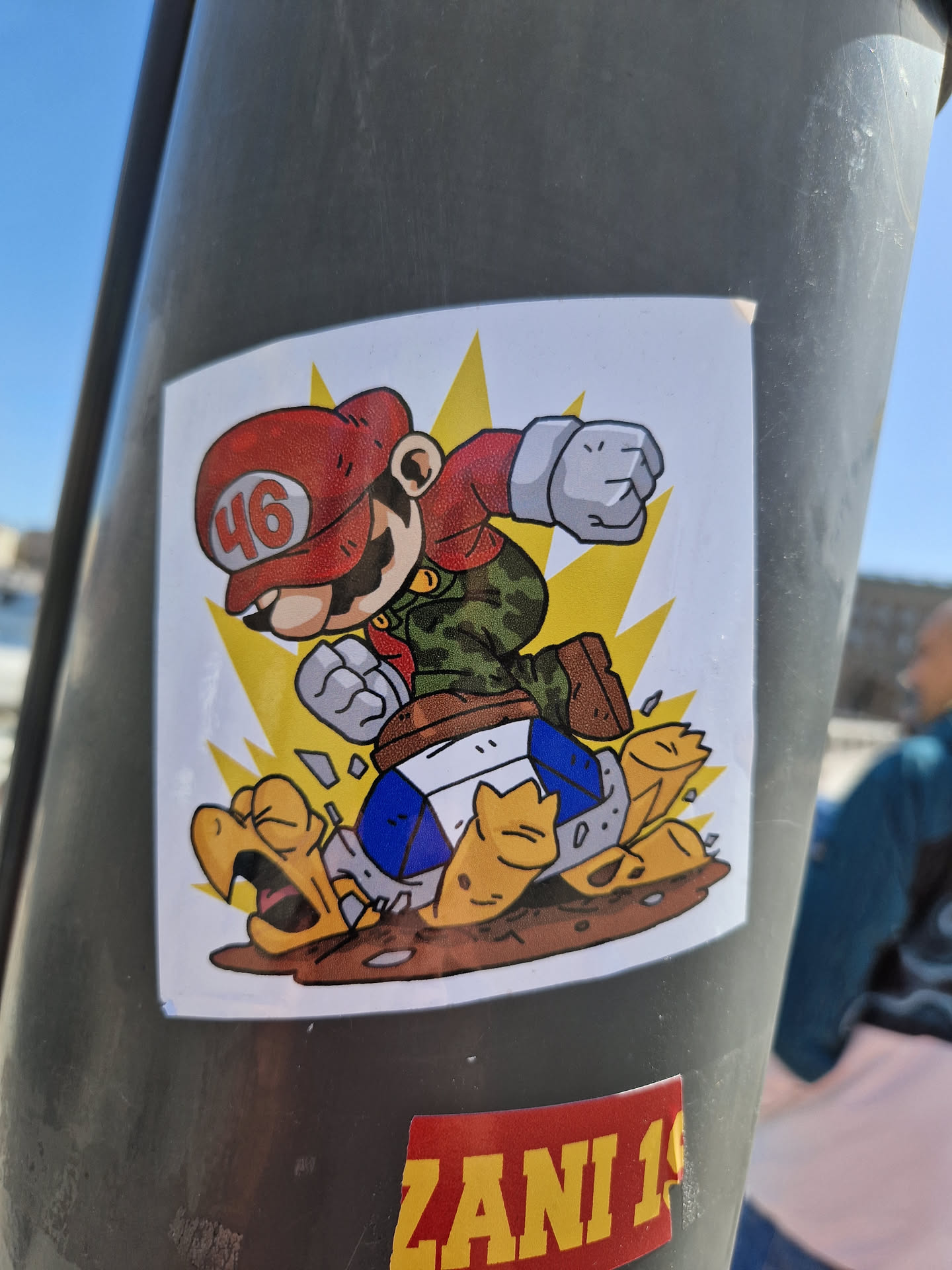 This sticker shows a stylized character resembling Mario from the Super Mario series, wearing a red cap with the number 46, kicking a Koopa Troopa. The art style is cartoonish and vibrant. The background is a simple yellow burst. The sticker is affixed to a gray pole.