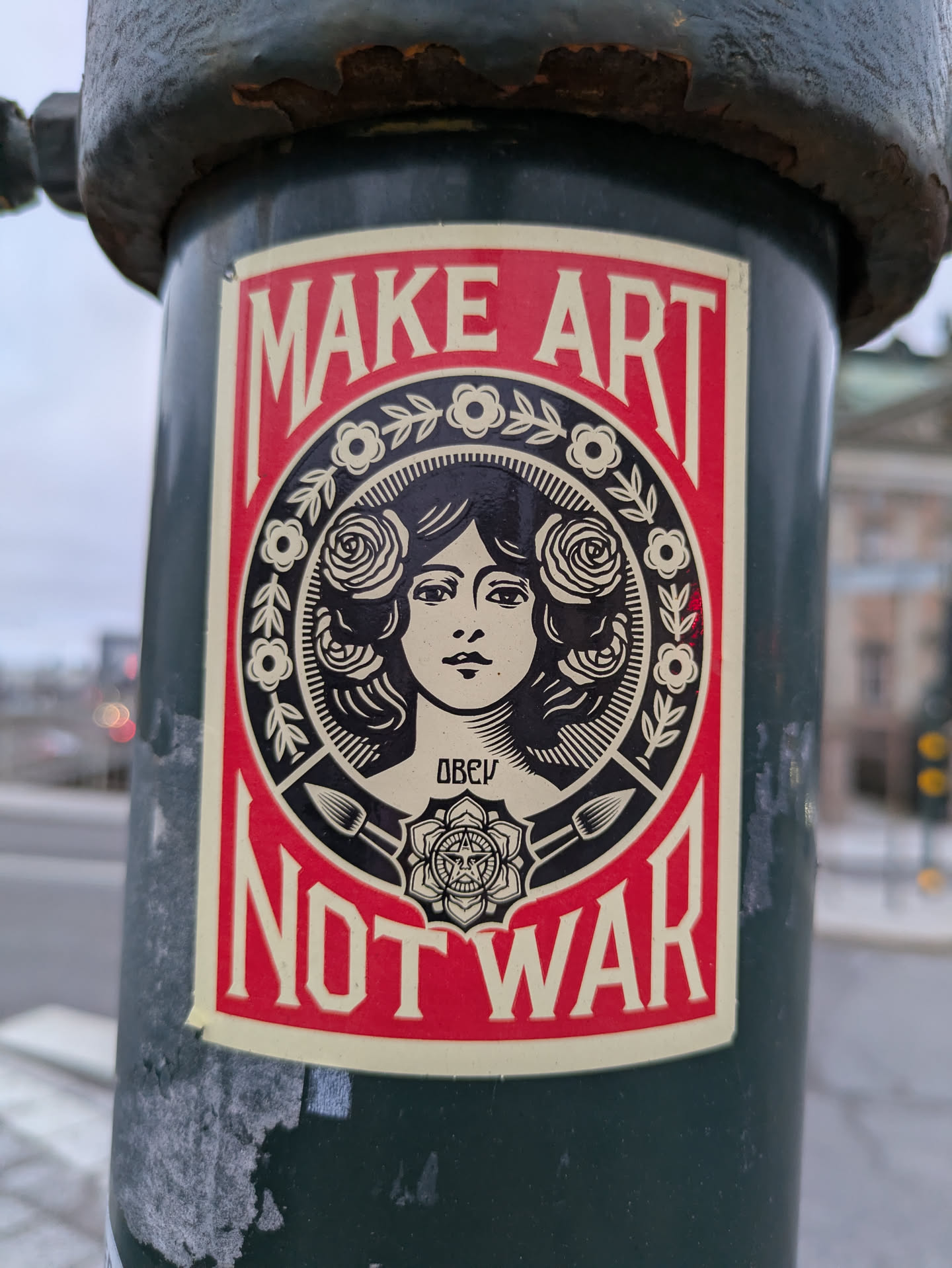 A sticker featuring a woman's face surrounded by roses and the text 'MAKE ART NOT WAR'. The sticker is predominantly red and beige, and the woman's face is styled in a vintage woodcut fashion. The OBEY logo is visible in the bottom center.