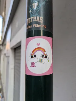 This is a round sticker on a lamppost. It features two cloud characters, each with large eyes and smiling faces. A colorful rainbow arcs between them, topped with a small pink heart. The cloud on the left is wearing pink cowboy boots, and the one on the right holds a small map.