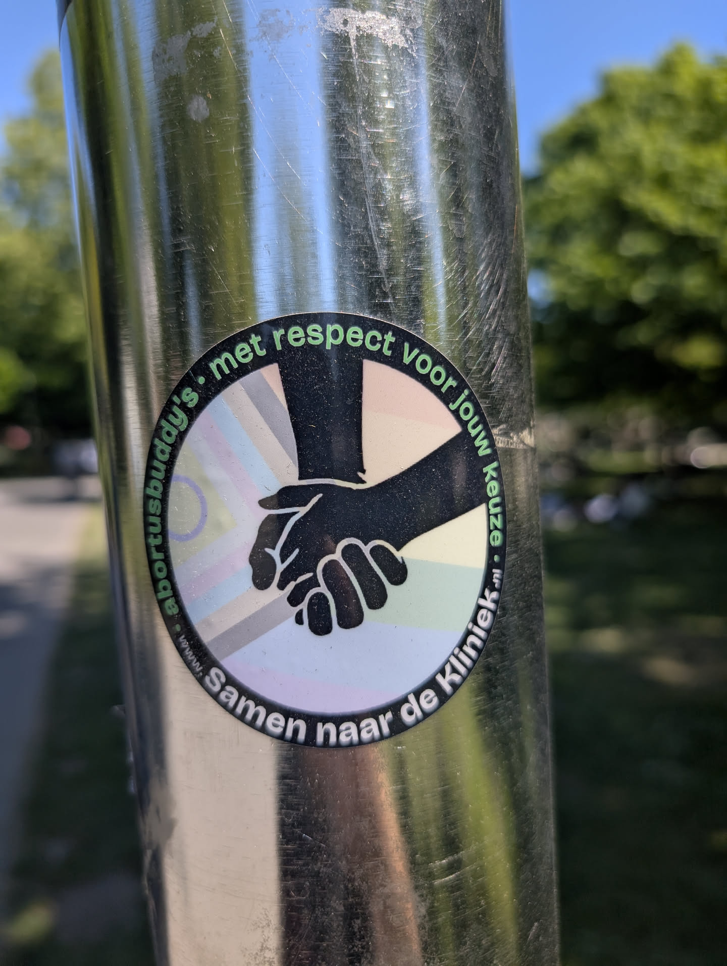 The sticker on the pole features two hands clasped together in a supportive gesture. The text 'met respect voor jouw keuze' (with respect for your choice) arcs above the hands, and 'Samen naar de Kliniek' (Together to the Clinic) arcs below. The sticker promotes respect for personal choice and suggests support for people visiting clinics.