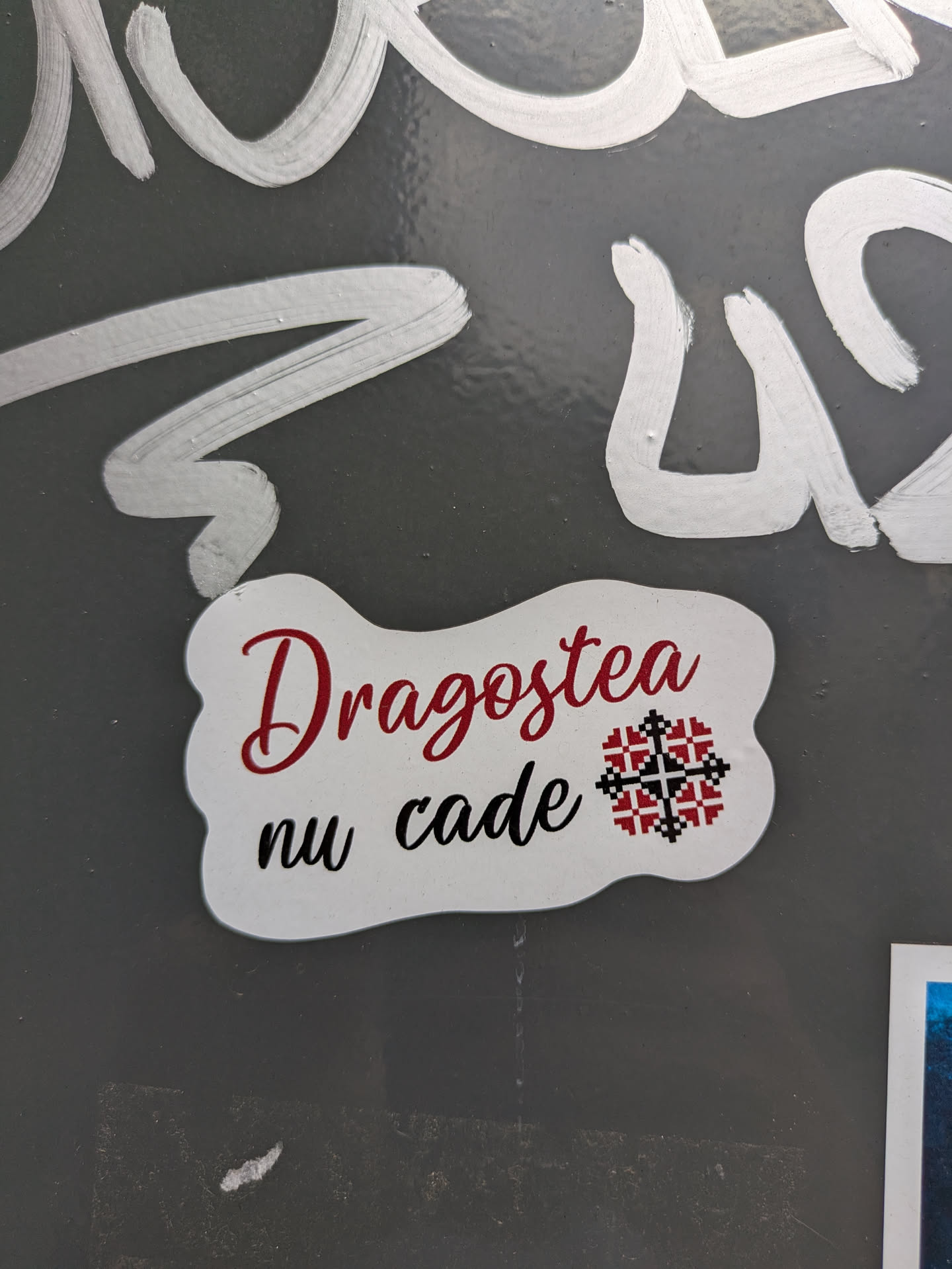 A white sticker with rounded, irregular edges. The text "Dragostea nu cade" is printed on the sticker in a mix of red and black fonts. A traditional Romanian design element, resembling a geometric flower, is included as part of the design.