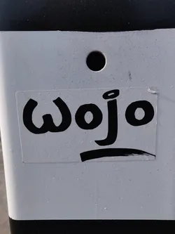 A white sticker with black text saying "Wojo" in a stylized font. The sticker is on a white pole with black stripes.
