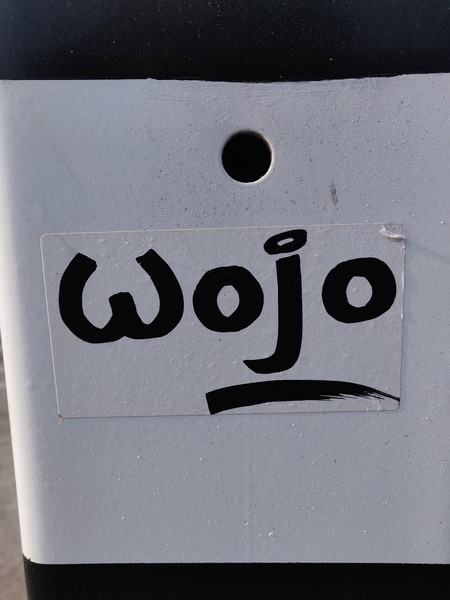 A white sticker with black text saying "Wojo" in a stylized font. The sticker is on a white pole with black stripes.