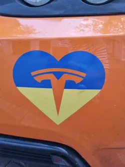 A heart-shaped sticker featuring the Tesla logo on a blue and yellow background, representing the Ukrainian flag.  The sticker is affixed to an orange vehicle.