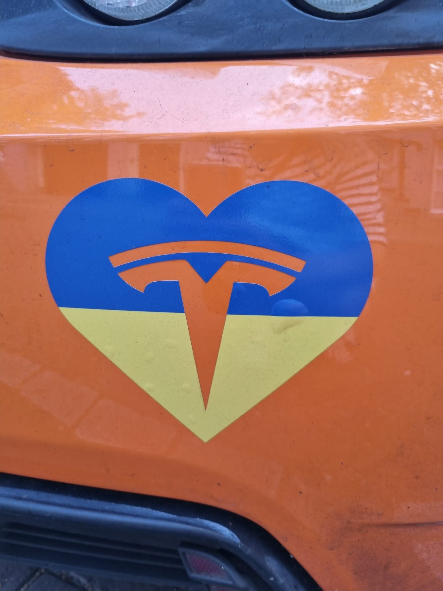 A heart-shaped sticker featuring the Tesla logo on a blue and yellow background, representing the Ukrainian flag.  The sticker is affixed to an orange vehicle.