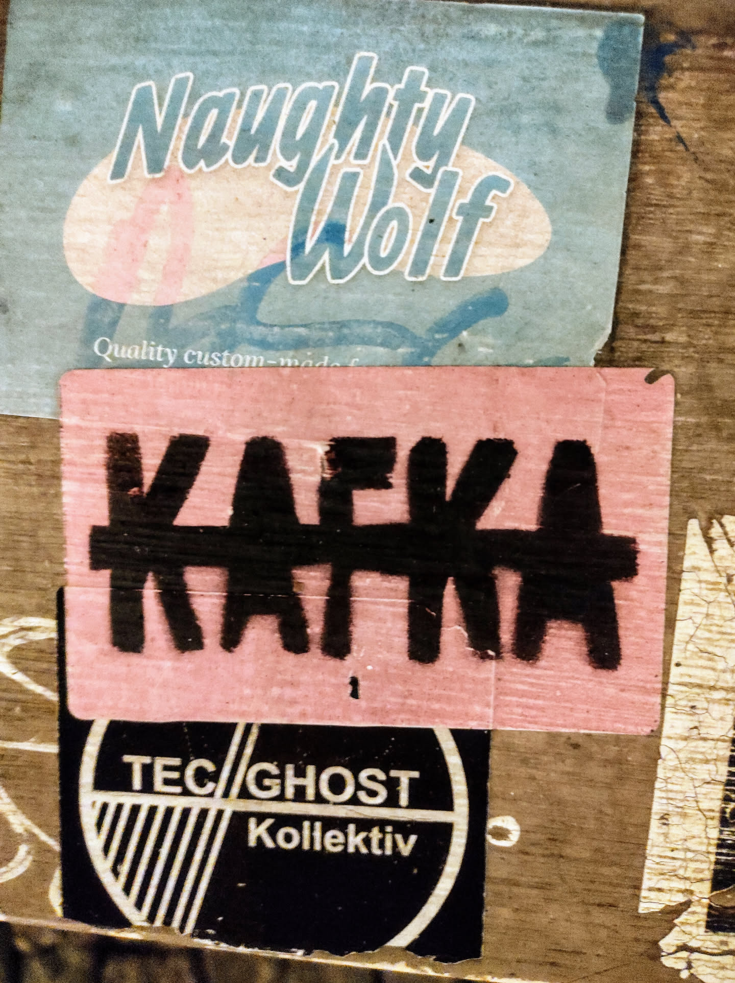 A close-up shot of a pink sticker with the word "Kafka" written in bold, black letters.  The sticker is slightly worn and appears to be pasted on a wooden surface with other stickers. One sticker above is a light blue sticker that says "Naughty Wolf" and one below says "TEC/GHOST Kollektiv".