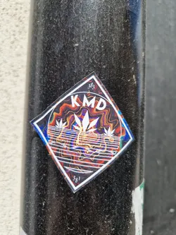 A diamond-shaped sticker with the letters KMD in the center. The background is a vibrant, swirling pattern of orange, red, and blue. Three stylized cannabis leaves are featured below the text.  The numbers 383 and 581 are visible in the corners of the sticker.