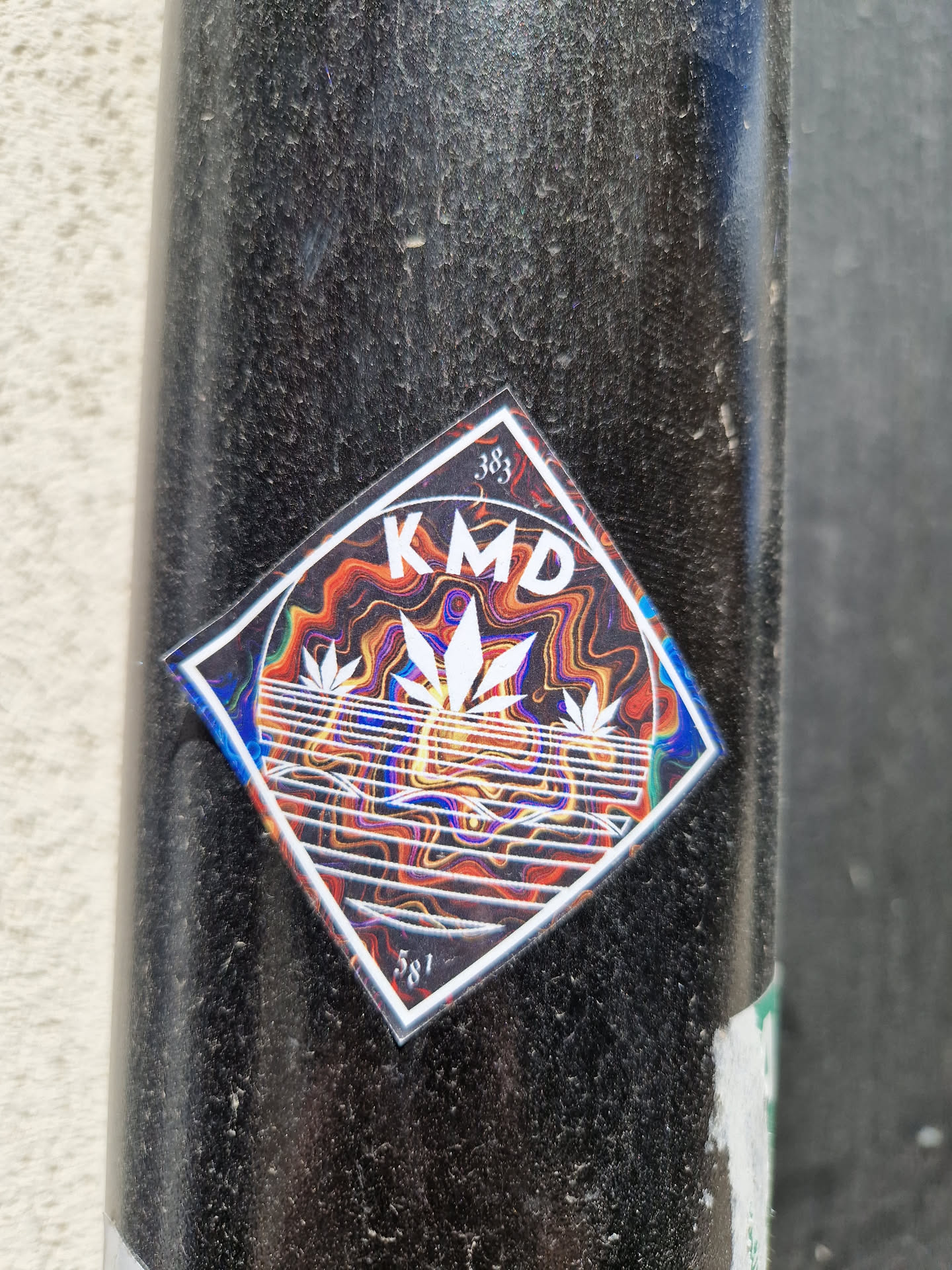 A diamond-shaped sticker with the letters KMD in the center. The background is a vibrant, swirling pattern of orange, red, and blue. Three stylized cannabis leaves are featured below the text.  The numbers 383 and 581 are visible in the corners of the sticker.