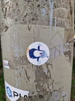 A cute round sticker featuring a cartoon penguin wearing a knitted hat.  The penguin is depicted swimming and has a happy expression. The sticker has a minimalist, slightly whimsical style. It is affixed to a weathered gray pole with other stickers and markings.