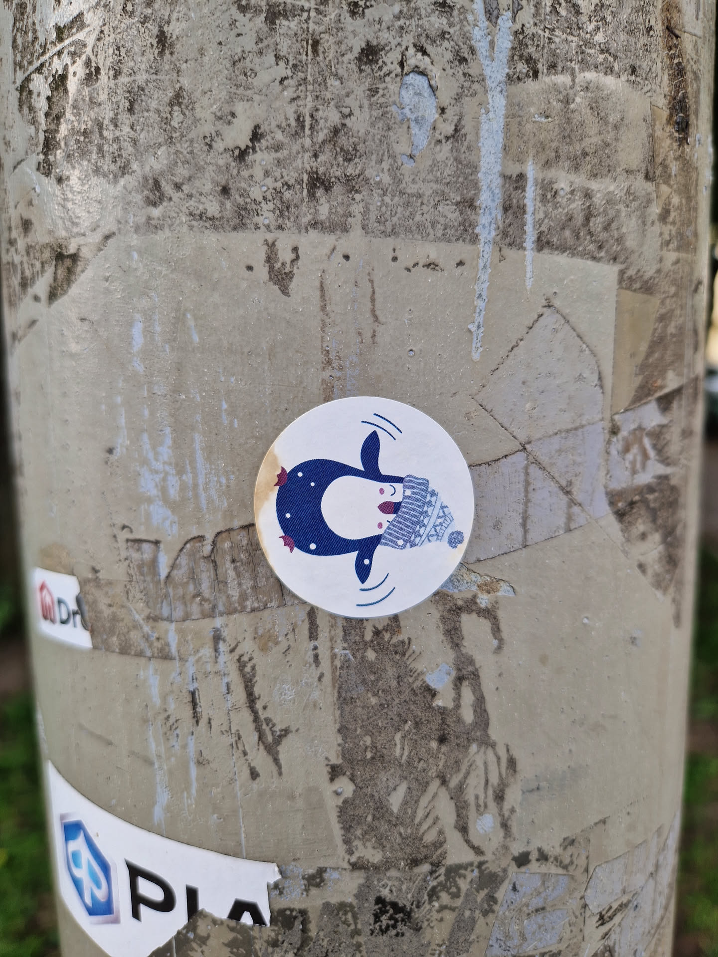 A cute round sticker featuring a cartoon penguin wearing a knitted hat.  The penguin is depicted swimming and has a happy expression. The sticker has a minimalist, slightly whimsical style. It is affixed to a weathered gray pole with other stickers and markings.