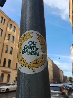 A round sticker featuring a laurel wreath design with the text "Ol' Dirty Bajen" in the center. The sticker is affixed to a metal pole, with buildings visible in the background.
