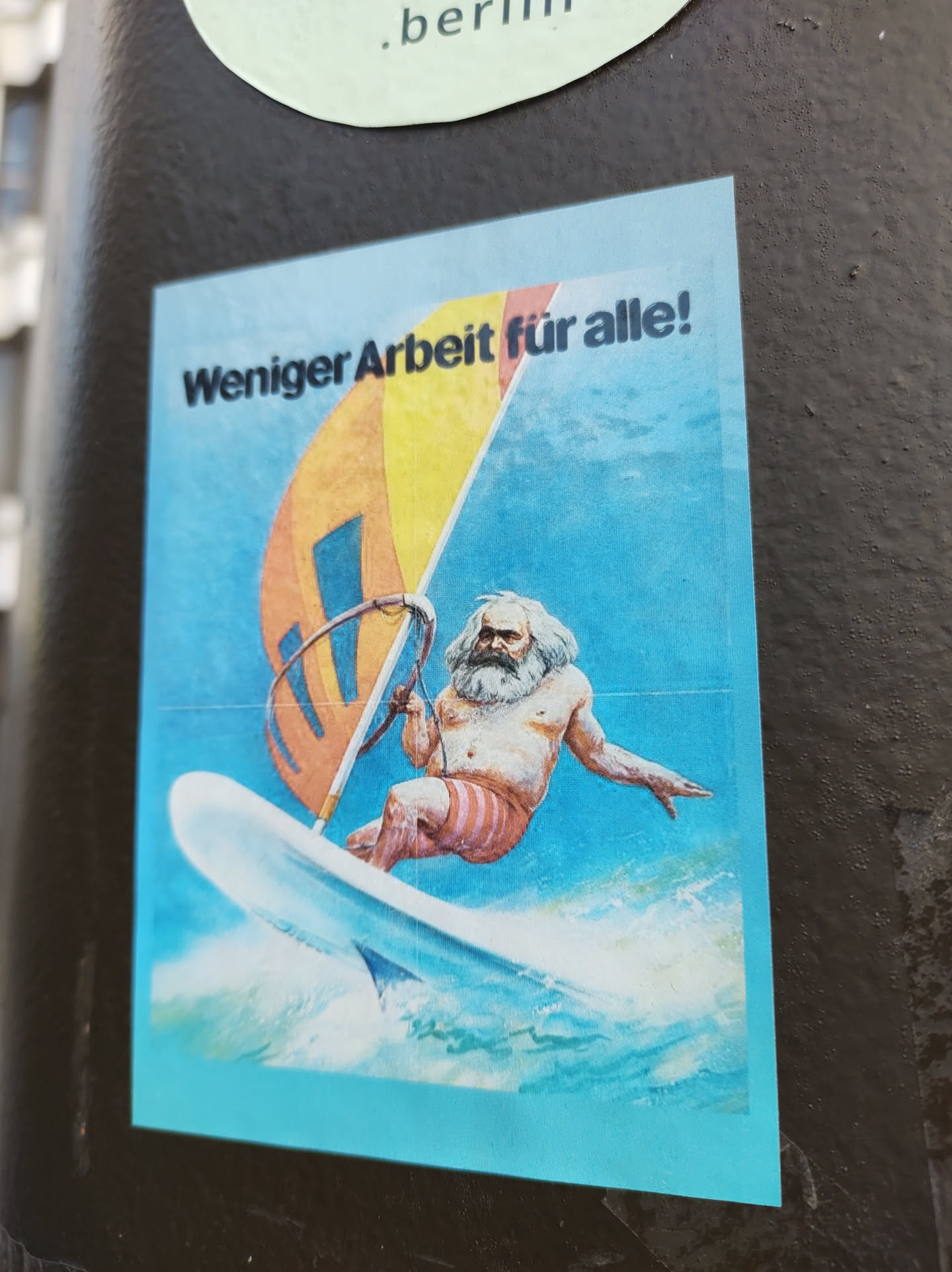 A sticker featuring a caricature of Karl Marx windsurfing with the text 'Weniger Arbeit für alle!' (Less work for all!)