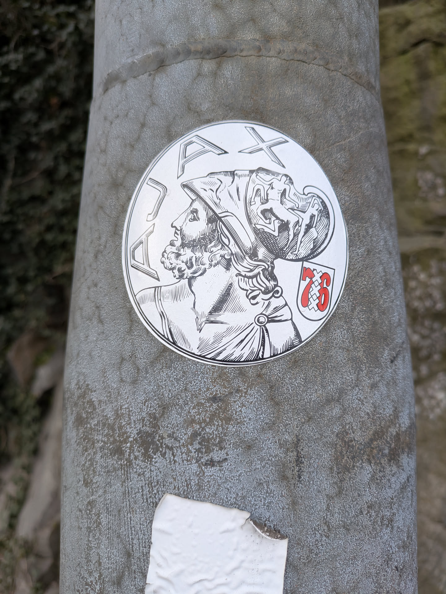 This sticker depicts a man with a beard and a helmet, drawn in a classical style. Above him are the letters 'DAX'. To his right is a shield with the number '76' on it, all within a round frame. The background is a neutral color of the pole where it's sticked.