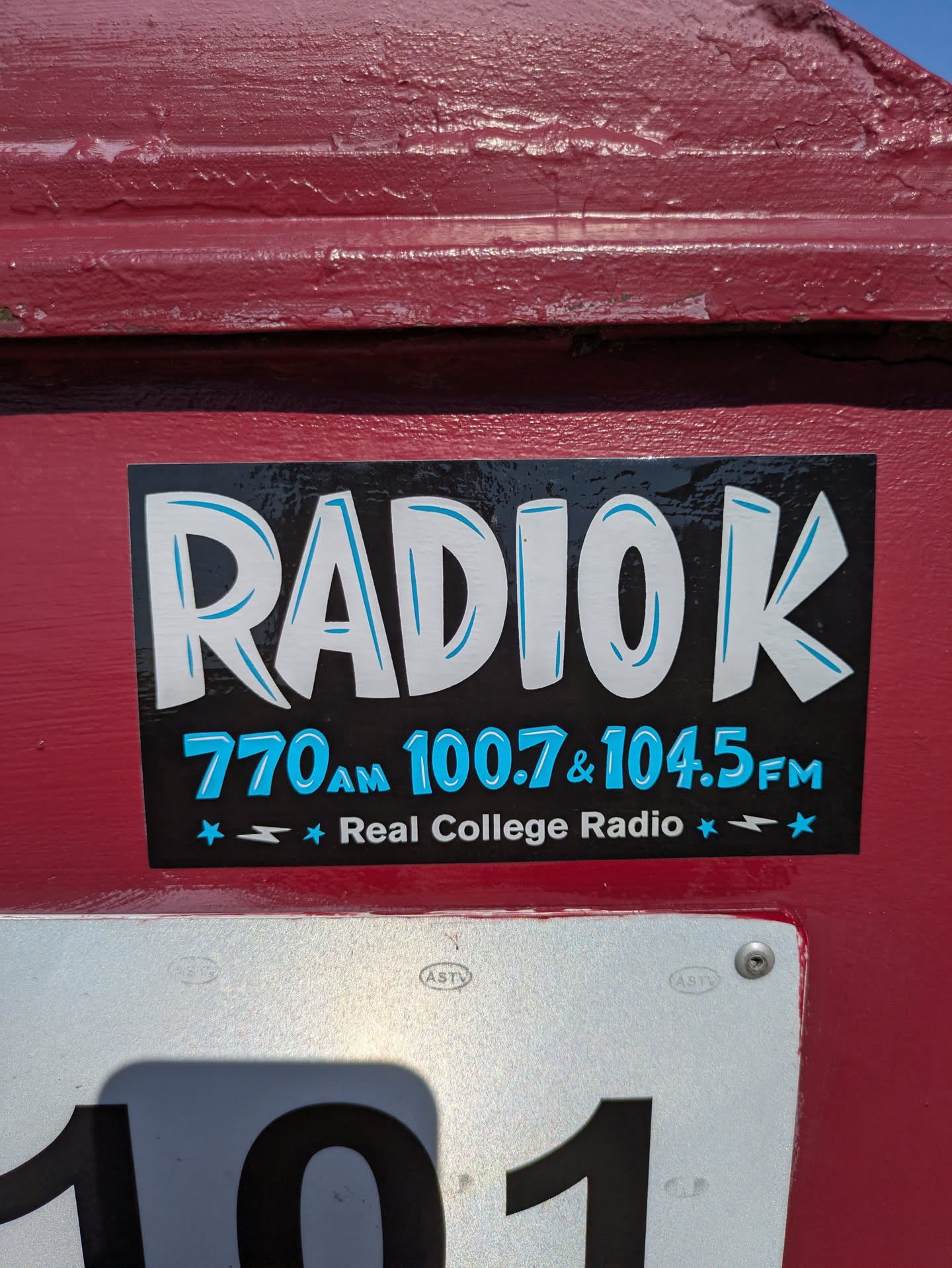 The sticker is rectangular with a black background that appears to have a slightly distressed or textured finish. The main text 'RADIO K' is displayed prominently in large, bold white letters with light blue outlines, giving them a playful, slightly bubbly appearance. Below the main logo, in smaller light blue text, are the frequencies '770AM 100.7 & 104.5FM'. At the bottom, in white text, it reads 'Real College Radio', flanked by small light blue stars and lightning bolt symbols. The sticker is affixed to a textured, dark red painted surface.