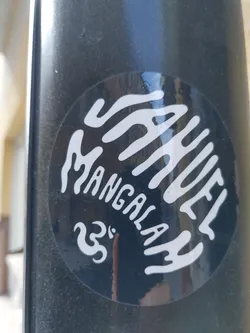 A black and white circular sticker with the words "MANUEL MANGALA" written in a stylized font. There's also a small symbol that looks like a stylized number 33. The sticker has a slightly glossy finish and is adhered to a dark surface.