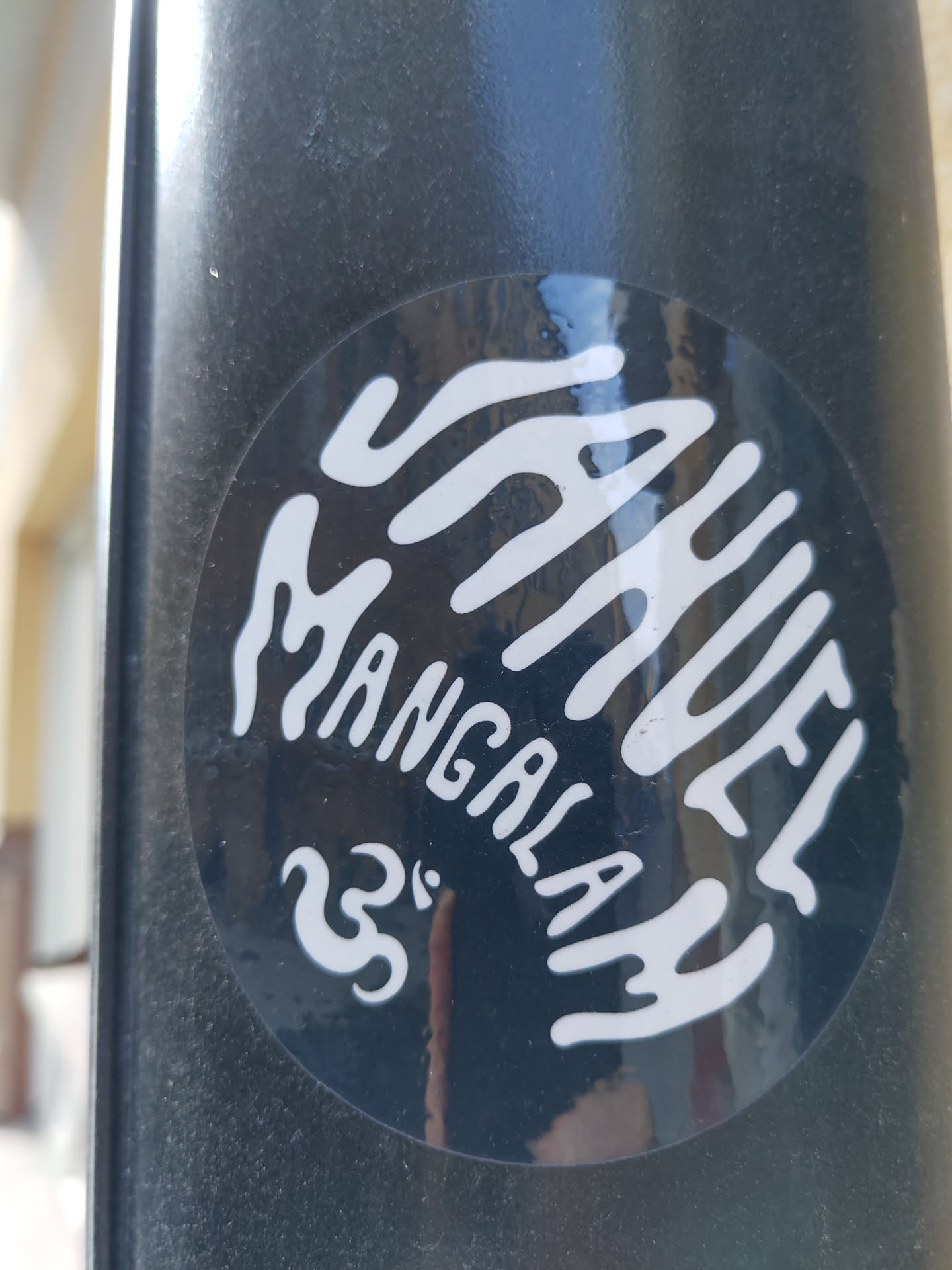 A black and white circular sticker with the words "MANUEL MANGALA" written in a stylized font. There's also a small symbol that looks like a stylized number 33. The sticker has a slightly glossy finish and is adhered to a dark surface.