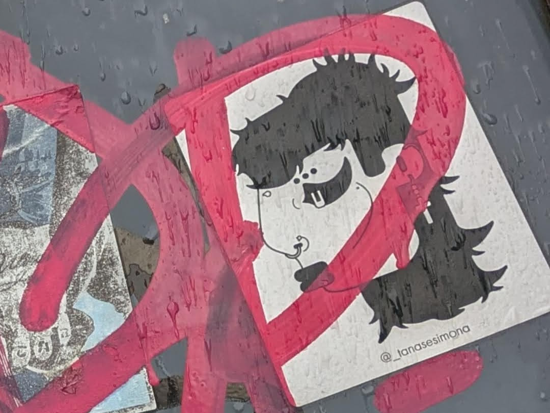 The sticker is predominantly white with a black graphic of a cartoonish face. The face has spiky black hair, large eyes with pupils, a small nose, and prominent buck teeth. A circular earring or piercing is visible on the nose. The sticker is overlaid with thick, bright pink paint strokes that create a dynamic, almost abstract pattern, partially obscuring the character. The overall impression is one of urban art or street art. The sticker appears to be slightly damp, with small water droplets visible on its surface and the surrounding area.