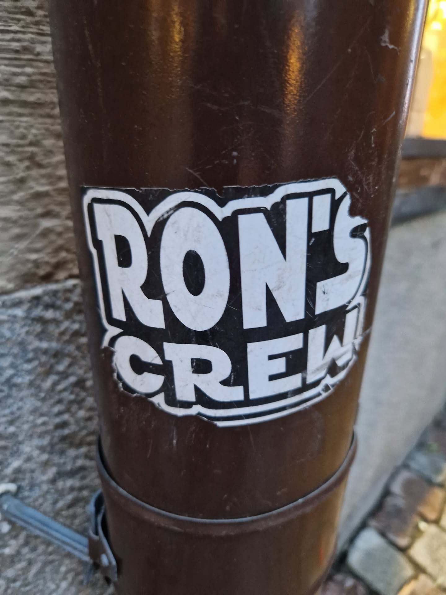 A worn sticker featuring the bold text 'RON'S CREW' in a stylized font. The sticker is affixed to a brown cylindrical object, possibly a drainpipe, outdoors.  The sticker shows some signs of wear and tear, adding to its street aesthetic.