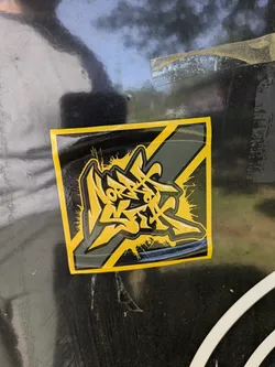 The image features a sticker with a black and yellow color scheme. The design is abstract, resembling stylized graffiti or an artistic logo. The sticker has a glossy finish and is affixed to a black surface with some wear and tear visible. The design incorporates sharp angles and what appear to be dynamic strokes, suggesting movement or energy.