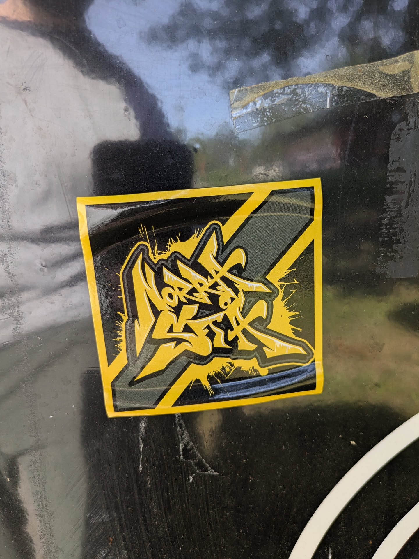 The image features a sticker with a black and yellow color scheme. The design is abstract, resembling stylized graffiti or an artistic logo. The sticker has a glossy finish and is affixed to a black surface with some wear and tear visible. The design incorporates sharp angles and what appear to be dynamic strokes, suggesting movement or energy.