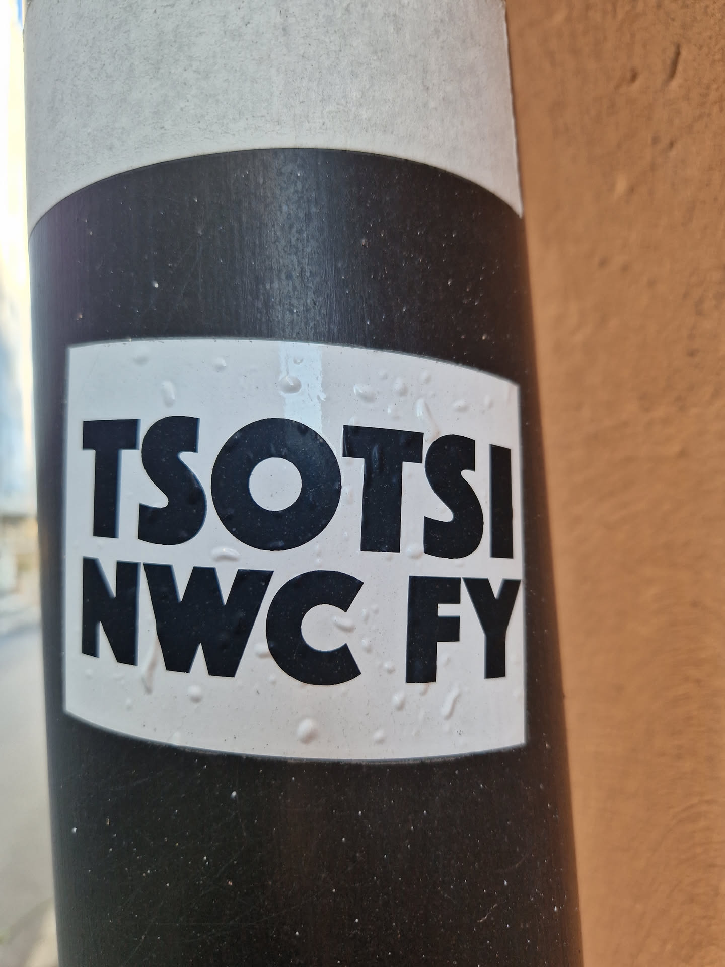 A black and white sticker with the text "TSOTSI NWC FY" in a bold, sans-serif font. The sticker is affixed to a dark gray pole.  The sticker shows some minor water droplets.