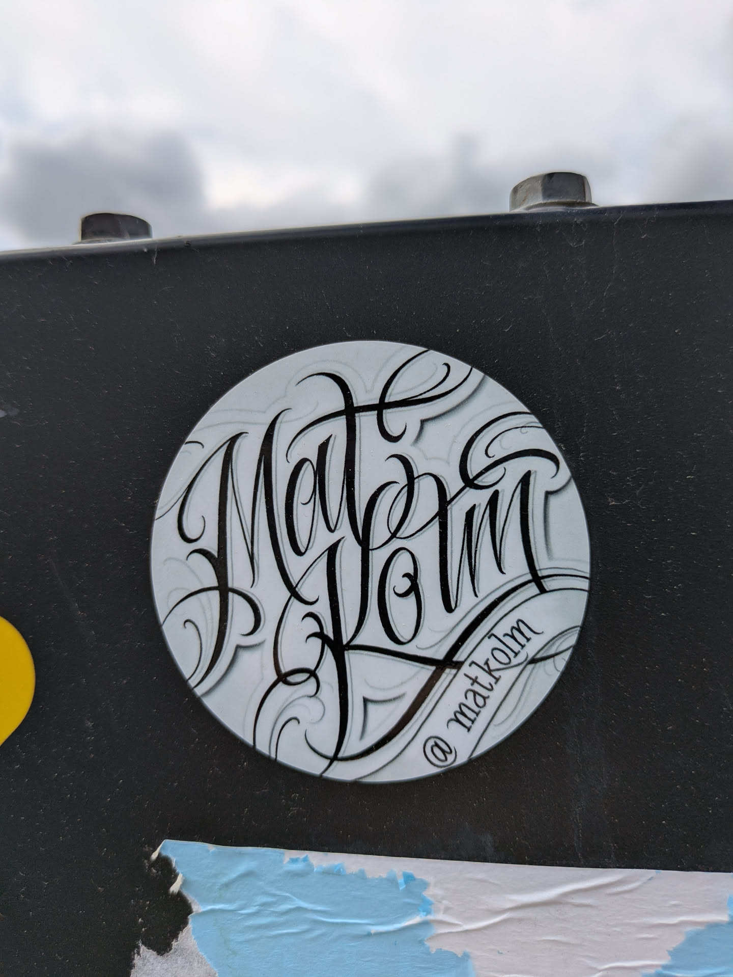 A round sticker featuring stylized black lettering on a white background with grey shadows. The text reads "Mat Kolm". The sticker is attached to a black metal surface, and part of other stickers are seen on the same surface.